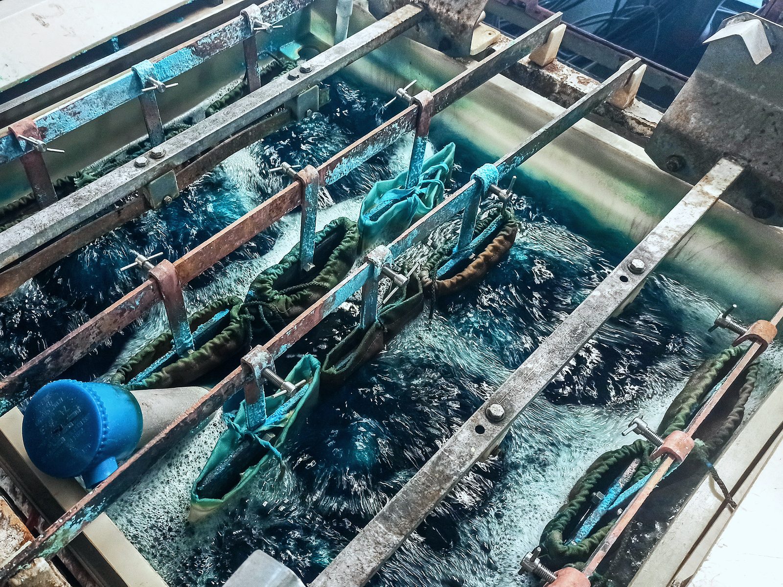 Metal parts in mesh bags hanging from rack bars, submerged in an agitated blue plating solution